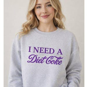 Könnte beinhalten: Hellgraues Sweatshirt mit dem lila Schriftzug "I NEED A Diet Coke". Der Text ist in einer Schreibschrift gehalten. Das Sweatshirt hat einen Rundhalsausschnitt und scheint aus einem weichen Material zu bestehen.