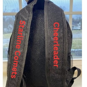 May include: Black backpack with glittery accents. The shoulder straps feature the words "Starline Comets" and "Cheerleader" in red lettering. The backpack has a mesh side pocket and adjustable straps.