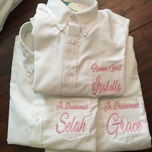 May include: White button-down shirts with embroidered pockets. One shirt reads "Flower Girl Isabella" in pink script. Two other shirts say "Jr. Bridesmaid" with the names "Selah" and "Grace" in pink script.