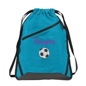 Può includere: Uno zaino a sacca blu brillante con spallacci neri e una base grigio scuro. La parte anteriore mostra il nome Harper in corsivo viola sopra la grafica di un pallone da calcio bianco e nero, il tutto racchiuso in un sottile riquadro.