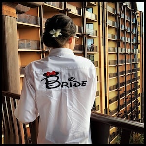 May include: A white button-down shirt with the word "BRIDE" embroidered on the back in black with a red bow and Mickey Mouse ears above the word.