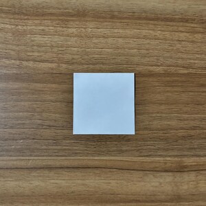 May include: A square, white sticky note centered on a wooden surface. The note is clean and blank, ready for use. The wood grain background is a warm brown.