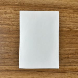 May include: A blank, rectangular white paper sheet, centered on a wooden surface. The paper is smooth and clean, with a bright white color. The wood grain of the surface is visible, with a warm, brown tone.