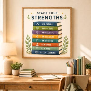 May include: A framed print with the words "STACK YOUR STRENGTHS" and a stack of books, each labeled with a positive affirmation. The books are in various colors, including blue, green, purple, and orange. The print is on a desk with books and a lamp.