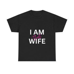 Puede incluir: Camiseta negra con el texto "I AM That WIFE" en letras blancas y rosas. El texto está centrado en la parte delantera de la camiseta. La camiseta tiene cuello redondo y parece estar hecha de un material suave.