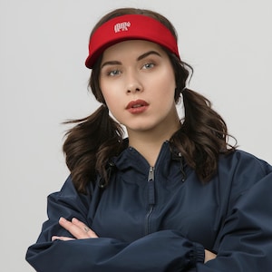 May include: A red visor with a white embroidered elephant design. The visor is made of a smooth material and has a curved brim. The visor is worn by a person wearing a navy blue jacket.