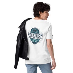 Op de afbeelding: Wit t-shirt met een teal en zwart grafisch ontwerp met de tekst "PRECISION ISN'T OPTIONAL". Het ontwerp is circulair en bevat elementen van een klok en een oog. Een zwarte leren jas is over de schouder gedrapeerd.