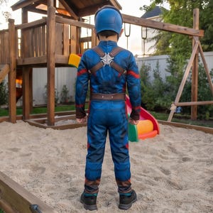 May include: A child wearing a Captain America costume, including a blue jumpsuit, helmet, and brown harness. The costume has red and brown accents. The child is standing in a sandbox near a wooden playset.