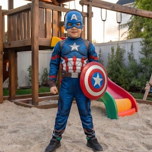 May include: A child wearing a Captain America costume, including a blue mask with a white "A", a blue and red suit, and a shield. The costume has a star on the chest and a brown belt. The child is standing in a sandbox near a wooden playset.