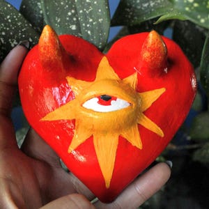 Könnte beinhalten: Eine rote herzförmige Skulptur mit orangefarbenen Hörnern und einem gelben Sternmuster mit einem Auge. Die Skulptur wird in einer Hand gehalten, mit grünem Laub im Hintergrund. Das Herz ist etwa 10 cm breit.