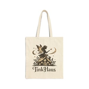 May include: Cream-colored tote bag featuring a brown illustration of a fairy on a house with a crescent moon and stars. The text "TinkHaus" is printed below the illustration. The bag has two cream-colored handles.