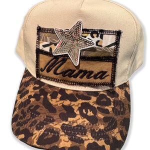 May include: A beige baseball cap with a leopard print brim. The cap features a patch with the word "Mama" and a rhinestone star. The patch is sewn onto the cap with black thread.