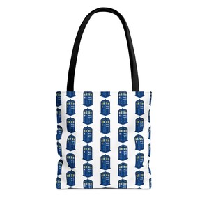 May include: A white tote bag with black shoulder straps featuring a repeating pattern of blue police box illustrations. Each blue box includes a small sign with the text Bad Wolf printed in a dark blue font on the side of the structure.