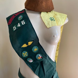 Puede incluir: Una banda verde de Girl Scout con un sombrero de fieltro marr&oacute;n, un pa&ntilde;uelo amarillo con un tr&eacute;bol verde y varias insignias bordadas. La banda tiene el n&uacute;mero "546" y las palabras "Northern Oakland" en ella.