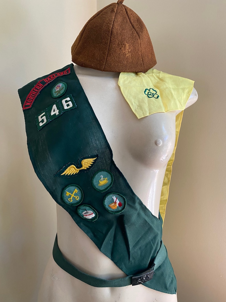 Puede incluir: Una banda verde de Girl Scout con un sombrero de fieltro marr&oacute;n, un pa&ntilde;uelo amarillo con un tr&eacute;bol verde y varias insignias bordadas. La banda tiene el n&uacute;mero "546" y las palabras "Northern Oakland" en ella.