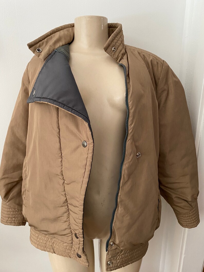Pode incluir: Uma jaqueta puffer marrom com z&iacute;per e fecho de press&atilde;o. A jaqueta tem gola alta e um design acolchoado.