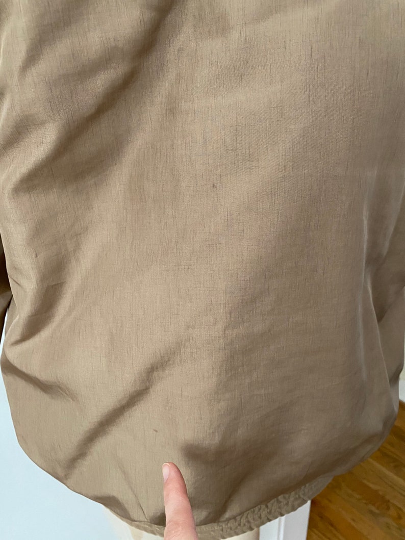 Pode incluir: Uma jaqueta bege com um acabamento liso e ligeiramente brilhante. A jaqueta tem um fecho de correr e uma gola alta.