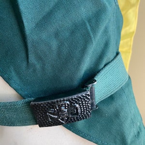 Puede incluir: Una faja de tela verde con una hebilla de metal negro con un emblema de scout. La hebilla est&aacute; asegurada a un cintur&oacute;n de tela verde.