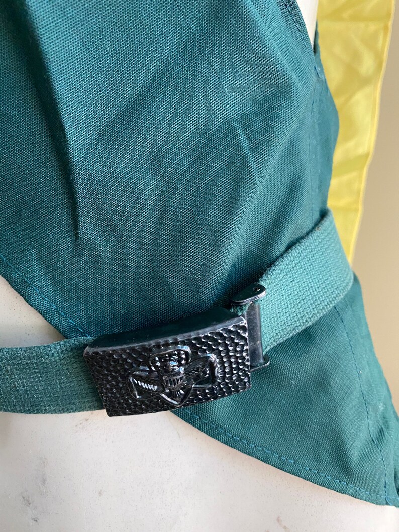 Puede incluir: Una faja de tela verde con una hebilla de metal negro con un emblema de scout. La hebilla est&aacute; asegurada a un cintur&oacute;n de tela verde.