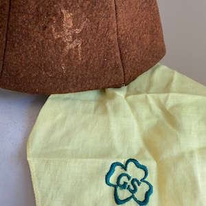 Puede incluir: Un sombrero de fieltro marr&oacute;n con un dise&ntilde;o bordado descolorido y una tela amarilla con una flor bordada verde que contiene las letras "GS".