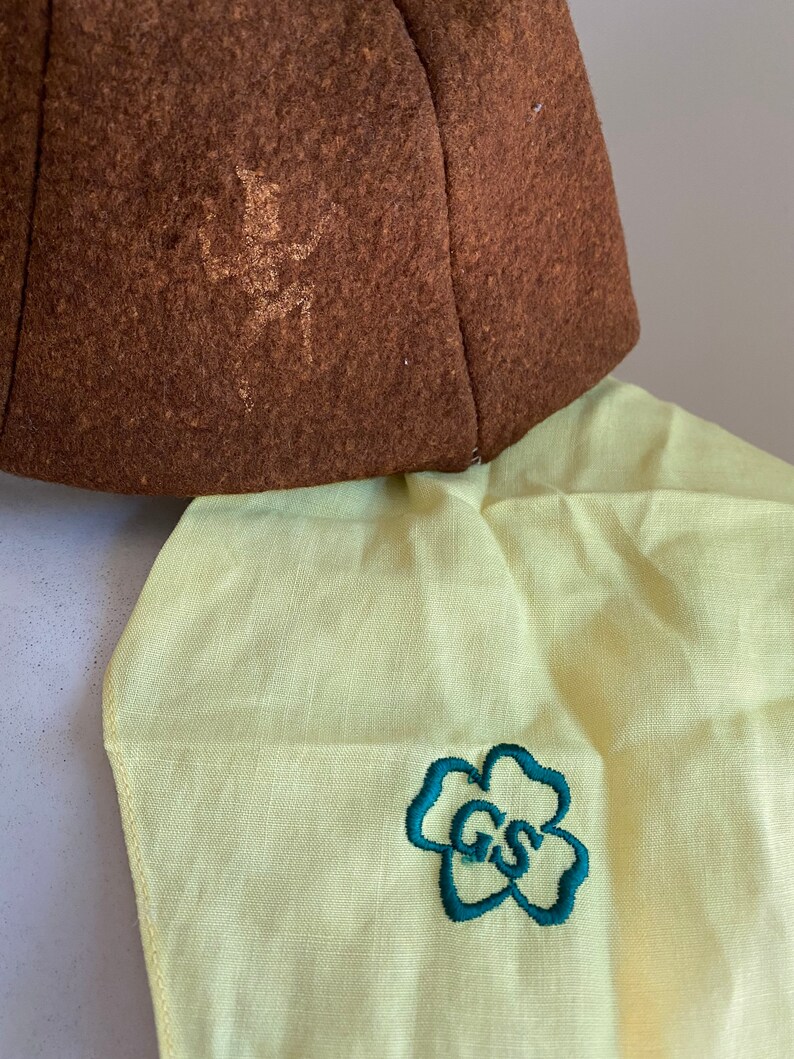 Puede incluir: Un sombrero de fieltro marr&oacute;n con un dise&ntilde;o bordado descolorido y una tela amarilla con una flor bordada verde que contiene las letras "GS".