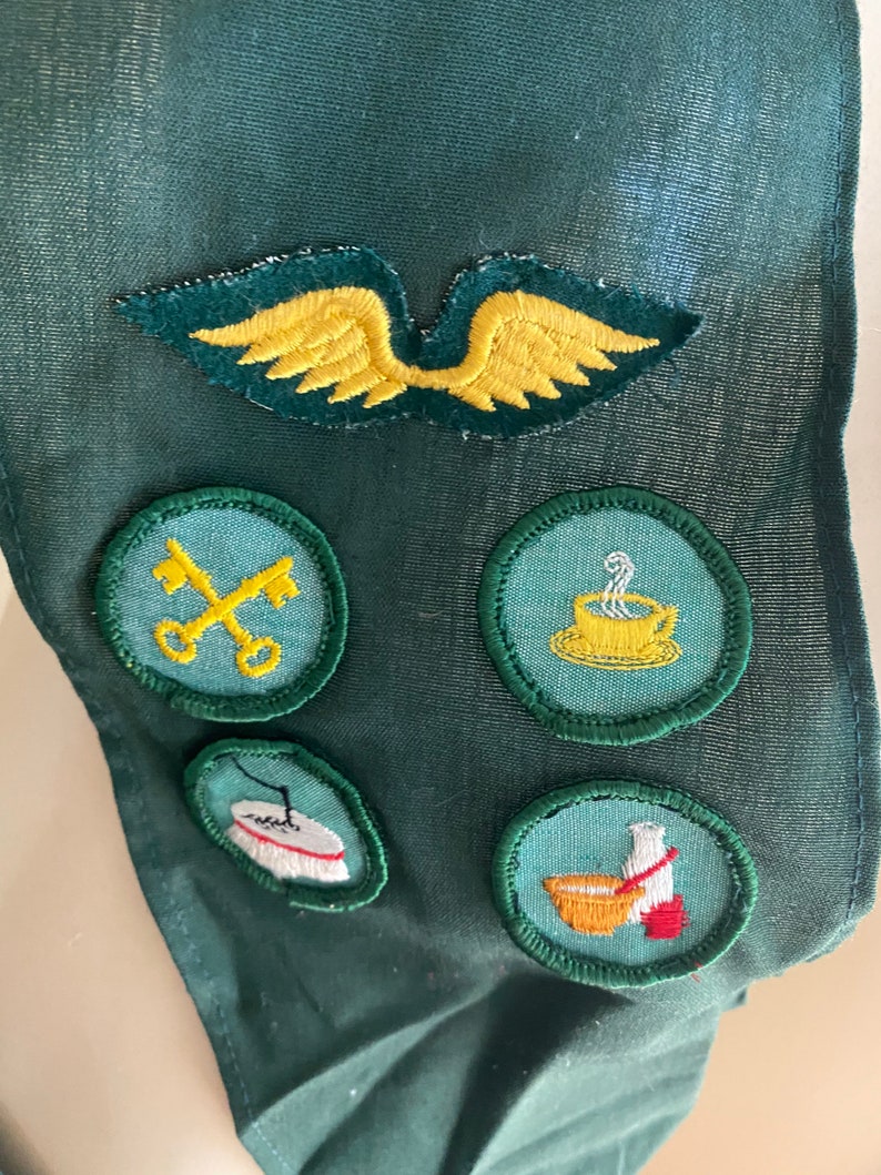 Puede incluir: Una faja de tela verde con cinco parches bordados. Los parches son amarillos, verdes y blancos con varios dise&ntilde;os, incluyendo una llave, una taza de caf&eacute;, una aguja de coser y un taz&oacute;n de sopa.