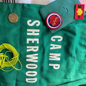 Puede incluir: Bolsa de lona verde con el texto "CAMP SHERWOOD" en blanco. La bolsa tiene una moneda marr&oacute;n, un parche bordado rojo, blanco y azul con el texto "SSS" y un parche bordado amarillo y rojo con una flecha negra.