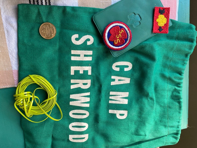 Puede incluir: Bolsa de lona verde con el texto "CAMP SHERWOOD" en blanco. La bolsa tiene una moneda marr&oacute;n, un parche bordado rojo, blanco y azul con el texto "SSS" y un parche bordado amarillo y rojo con una flecha negra.