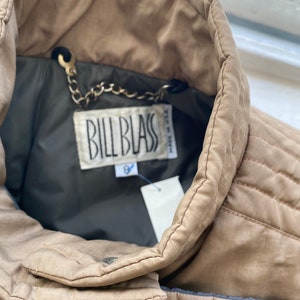 Pode incluir: Uma jaqueta puffer bege com uma etiqueta marrom Bill Blass e uma corrente prateada presa ao colarinho. A etiqueta diz "Made in Italy".