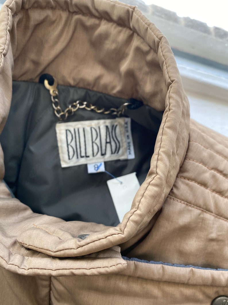 Pode incluir: Uma jaqueta puffer bege com uma etiqueta marrom Bill Blass e uma corrente prateada presa ao colarinho. A etiqueta diz "Made in Italy".