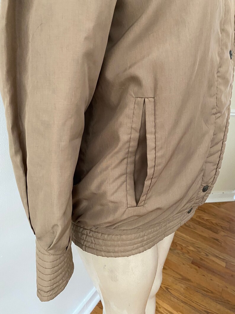 Pode incluir: Uma jaqueta marrom com fecho de z&iacute;per e um bolso do lado esquerdo. A jaqueta tem uma bainha e punhos canelados.