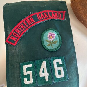 Puede incluir: Sash verde de Girl Scout con un parche rojo y blanco "Northern Oakland", un parche de flor verde y un parche de n&uacute;mero blanco "546".