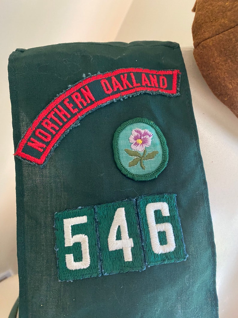 Puede incluir: Sash verde de Girl Scout con un parche rojo y blanco "Northern Oakland", un parche de flor verde y un parche de n&uacute;mero blanco "546".