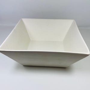 May include: A square, cream-coloured ceramic bowl with a slightly textured surface. The bowl has a simple, modern design with angled sides and a wide, open top. Suitable for serving salads or as a decorative item.