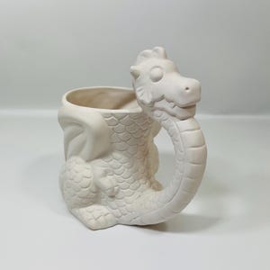 May include: An unpainted ceramic dragon mug. The mug is off-white and features a dragon head handle, wings, and a scaled body. The mug is designed for painting and customisation.