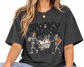 May include: A charcoal gray t-shirt featuring a graphic of three skeletons. One skeleton wears a hat and holds a coffee cup, one sits in a shopping cart, and one wears a beanie and holds a pumpkin. The text reads Spooky Season.