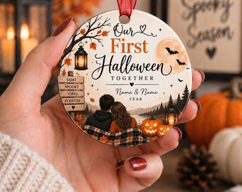 May include: A round ceramic ornament with a red ribbon featuring a printed illustration of a couple sitting under a tree with a lantern, pumpkins, and a full moon. The text reads Our First Halloween TOGETHER, Name & Name, YEAR, and SAME SPOOKY VIBES FOREVER.