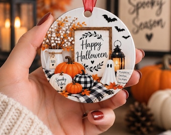 May include: A round ceramic ornament with a red ribbon hanger featuring a Halloween scene with pumpkins, a ghost, a lantern, and a sign that reads Happy Halloween. The design includes a white pitcher, a black and white plaid cloth, and bats.