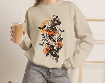 May include: A beige crewneck sweatshirt featuring a graphic print of a skeletal hand holding a glass bottle filled with orange liquid, surrounded by a yellow crescent moon, black bats, and orange flowers with dark green leaves.