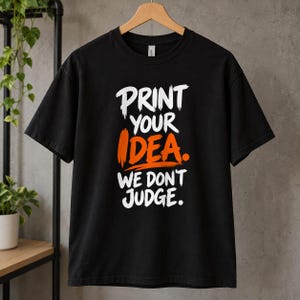 May include: A black short-sleeved t-shirt featuring the white and orange text "PRINT YOUR IDEA. WE DON'T JUDGE." The graphic is centered on the chest in a bold, brush-stroke style font. The shirt is displayed on a wooden hanger.