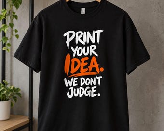 May include: A black short-sleeved t-shirt featuring the white and orange text "PRINT YOUR IDEA. WE DON'T JUDGE." The graphic is centered on the chest in a bold, brush-stroke style font. The shirt is displayed on a wooden hanger.