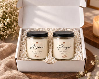 May include: Two glass candles with black lids inside a white cardboard gift box filled with white packing material. The left candle label reads Arjun, My Person Always. The right candle label reads Priya, My Happy Place. Both feature a small heart.