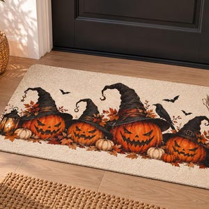 May include: A rectangular doormat with a beige background featuring a printed design of four orange jack-o'-lanterns wearing black witch hats, surrounded by autumn leaves, a lantern, a black crow, a broom, and a black cat.