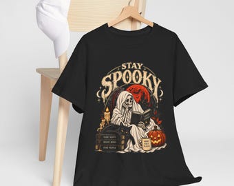 May include: A black t-shirt featuring a graphic of a skeleton reading a book titled Ghost Stories, with the text Stay Spooky, a black cat, a stack of books labeled Dark Nights Bright Minds Kind People, a pumpkin, and a mug saying More Boos Please.