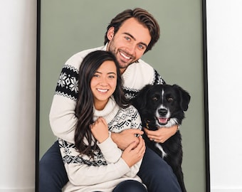 May include: A framed portrait featuring two individuals and a black and white dog. The individuals are wearing matching cream-colored sweaters with black geometric patterns. The dog has a white chest and a white stripe on its face.