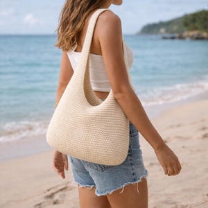 Puede incluir: Un bolso de hombro de crochet color crema, de forma redondeada y con una correa ancha. El bolso se lleva al hombro. El fondo muestra una playa con agua azul y un cielo despejado.