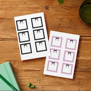 May include: Two sheets of stickers with a bow design. One sheet has black borders and bows, the other has pink borders and bows. A green pencil, notebook, and push pins are also on the wooden surface.