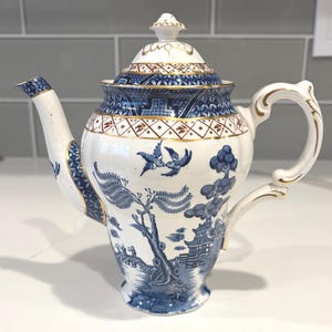 Può includere: Una teiera in ceramica bianca con un motivo blu a salice, caratterizzata da dettagliate illustrazioni paesaggistiche con uccelli, alberi e strutture architettoniche. La teiera presenta un coperchio coordinato, un beccuccio curvo e un manico dorato.