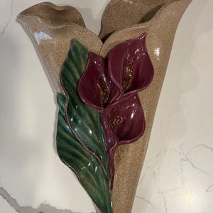 May include: A beige ceramic wall vase with a speckled finish, featuring three burgundy calla lilies and a green leaf. The vase has a unique, organic shape, perfect for displaying flowers or as a decorative piece. The vase measures approximately 12 inches in height.