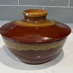 Puede incluir: Un cuenco de cerámica con tapa. El cuenco y la tapa son de color marrón rojizo oscuro, con una banda beige más clara alrededor del centro del cuenco. La tapa tiene un pequeño asa redondeada.